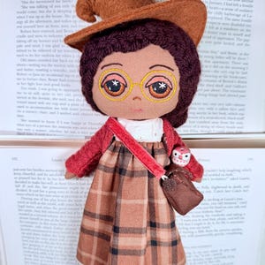 May include: A handmade doll with a brown witch hat, glasses, and a plaid dress. The doll has a red sweater and a small purse with an owl attached. The doll is sitting on top of an open book.