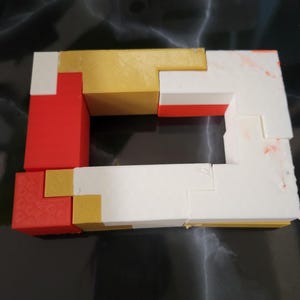 May include: A puzzle made of interlocking blocks in red, white, and gold. The blocks are arranged in a frame shape, with a hole in the center.