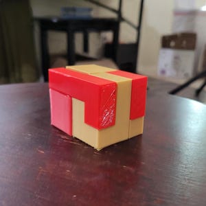 May include: A multi-colored puzzle cube with red, tan, and pink components. The cube is composed of several interlocking pieces, creating a three-dimensional shape. The puzzle is sitting on a dark wooden surface.