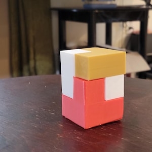 May include: A multi-colored puzzle cube composed of several interlocking blocks. The cube features a combination of red, white, and yellow-orange pieces, creating a visually engaging geometric design. The puzzle is displayed on a dark wooden surface.