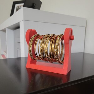 May include: A red wooden bangle holder displaying several gold bangles. The bangles are adorned with intricate patterns and sparkling embellishments. The holder is a vibrant red, providing a striking contrast to the gold jewelry.