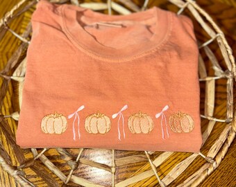 May include: An orange crew neck t-shirt with embroidered pumpkins and white bows. The shirt is folded and placed inside a woven basket. The pumpkins are a light orange color.