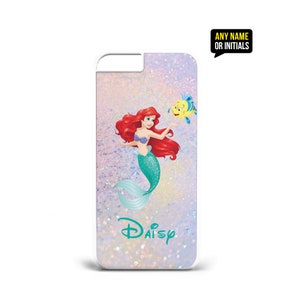 Op de afbeelding: Een witte iPhone-hoes met een glinsterende achtergrond waarop Ariel, de Kleine Zeemeermin, te zien is met haar rode haar en groene staart. Ze houdt een gele vis met blauwe vinnen vast. De tekst "Daisy" staat in het groen op de hoes.
