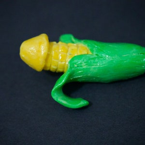 May include: A yellow and green corn cob sculpture. The corn is partially peeled back to reveal the kernels.