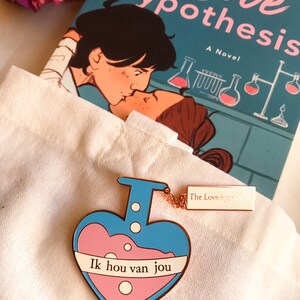 May include: A heart-shaped enamel pin in blue and pink, featuring the text "Ik hou van jou" and a small tag with "The Love Hypothesis". The novel "The Love Hypothesis" is visible in the background.