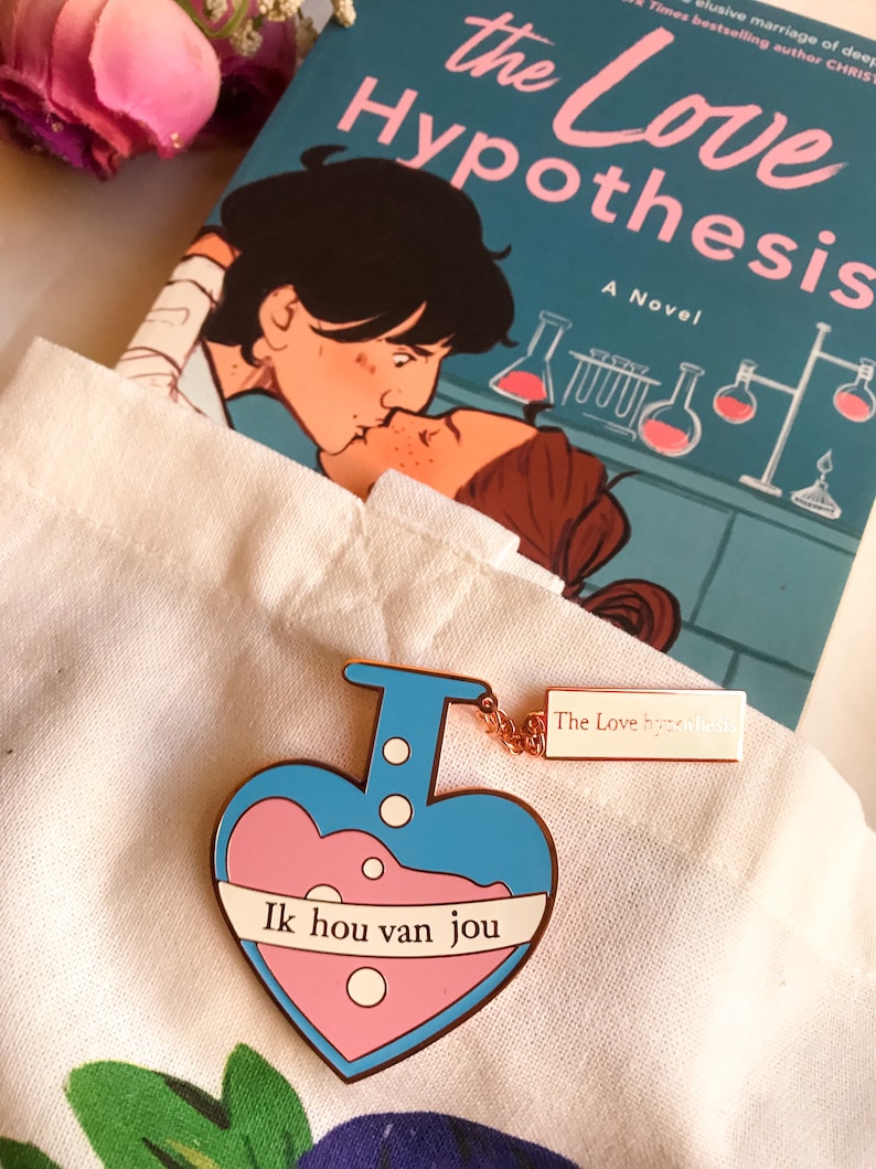 May include: A heart-shaped enamel pin in blue and pink, featuring the text "Ik hou van jou" and a small tag with "The Love Hypothesis". The novel "The Love Hypothesis" is visible in the background.