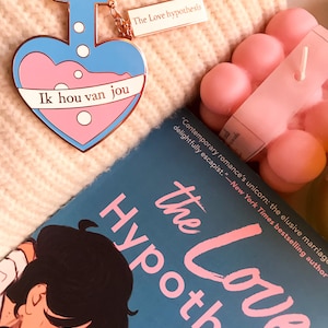 May include: A heart-shaped enamel pin with a blue and pink potion design and the text "Ik hou van jou." A rectangular tag reads "The Love hypothesis." A book titled "The Love Hypothesis" is also visible.