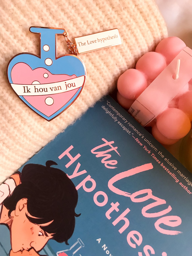 May include: A heart-shaped enamel pin with a blue and pink potion design and the text "Ik hou van jou." A rectangular tag reads "The Love hypothesis." A book titled "The Love Hypothesis" is also visible.