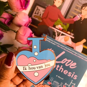 May include: A heart-shaped enamel pin with a blue outline and a pink liquid design. The pin features the text "Ik hou van jou" on a white banner. A book titled "The Love Hypothesis" is visible in the background.