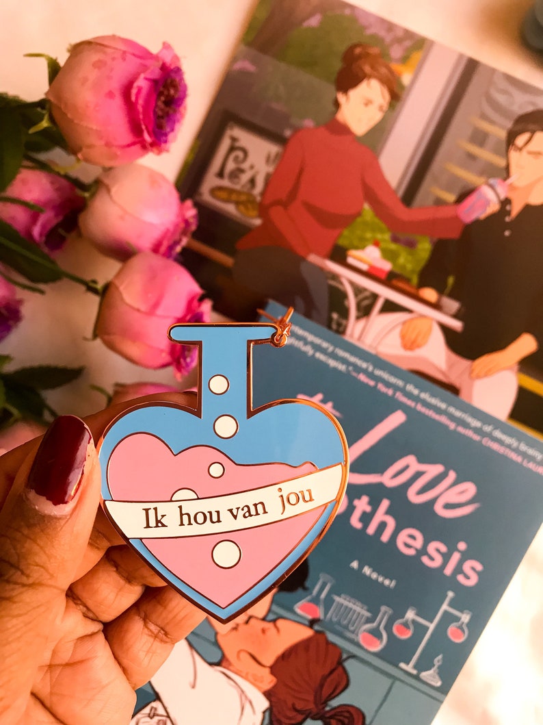 May include: A heart-shaped enamel pin with a blue outline and a pink liquid design. The pin features the text "Ik hou van jou" on a white banner. A book titled "The Love Hypothesis" is visible in the background.