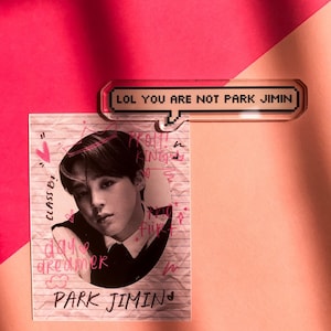 May include: A photo card featuring a black and white image of Park Jimin, with pink handwritten text and doodles. Above it, a clear acrylic pin with the text "LOL YOU ARE NOT PARK JIMIN" in pixelated font.