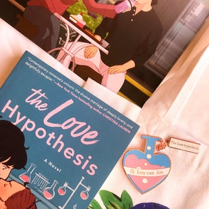 May include: A novel titled "The Love Hypothesis" with a teal cover and illustrations of lab equipment. A heart-shaped enamel pin with the text "Ik hou van jou" and a print of a couple are also visible.