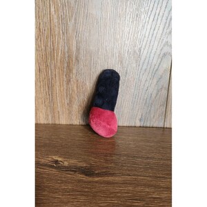Puede incluir: Un juguete de peluche con forma de barra de labios. La parte superior es negra y la inferior es de color rojo vibrante. El juguete está hecho de un material suave y difuso y está diseñado para parecerse a un producto cosmético.