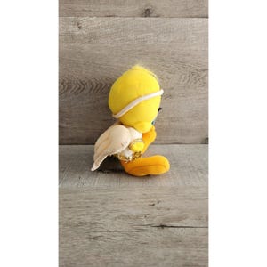 May include: A yellow plush Tweety Bird toy wearing a white angel costume with gold accents. The toy is sitting on a wooden surface.