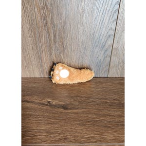 Puede incluir: Un juguete afelpado en forma de pata, de color marrón, con almohadillas blancas y garras azules. El juguete está sobre una superficie de madera, mostrando su textura suave y su diseño juguetón. Este artículo es probablemente un juguete para mascotas.