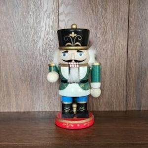 May include: A wooden nutcracker figurine featuring a black hat with gold detailing, a green and white jacket, blue trousers, and black boots. The figure has a white beard, painted facial features, and stands on a circular red base.