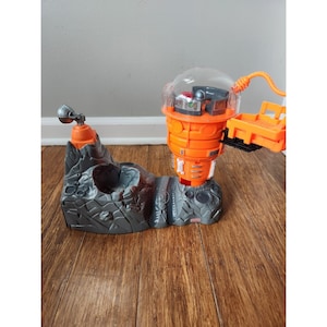 May include: A grey and orange toy playset featuring a rocket ship, a small vehicle, and a rocky terrain. The rocket ship has a clear dome and a small opening on the side. The vehicle has a small bed and a handle. The rocky terrain has a small opening on the side.