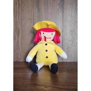 May include: A plush doll with a yellow coat, hat, and red hair. The doll has a simple face with a stitched smile and black button eyes. The doll's arms and legs are white and black, respectively. The doll is sitting on a wooden surface.