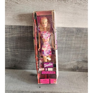 May include: A Barbie doll in a box. The doll has blonde hair and is wearing a pink and purple patterned outfit. The box has the words "Barbie HIP 2 SQUARE" printed on it.