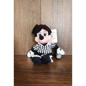 Könnte beinhalten: Eine Plüschpuppe von Mickey Mouse mit einem schwarz-weiß gestreiften Hemd, schwarzen Shorts und weißen Handschuhen. Die Puppe hat einen schwarzen Kopf und ein angebrachtes Etikett. Die Puppe sitzt auf einer Holzoberfläche.