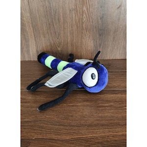 Puede incluir: Un insecto de peluche con cuerpo azul, abdomen a rayas verdes y negras y ojos blancos grandes. El juguete tiene alas grises y patas y antenas negras. El juguete está sobre una superficie de madera.