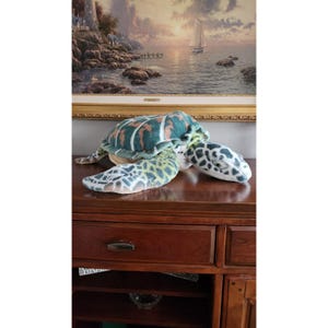 Puede incluir: Un gran juguete de peluche de tortuga marina con una concha verde, marrón y blanca. La tortuga está tumbada sobre una superficie de madera.