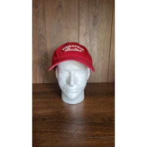 May include: A red baseball cap with the text "Universal Studios" in white script. The cap is displayed on a white mannequin head against a wooden background.