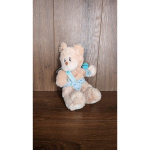 Puede incluir: Un oso de peluche sentado, con pelaje marrón claro y hocico blanco. Lleva un babero azul y un pañal con un estampado de pequeñas estrellas amarillas y puntos azules. El oso sostiene una sonaja azul.