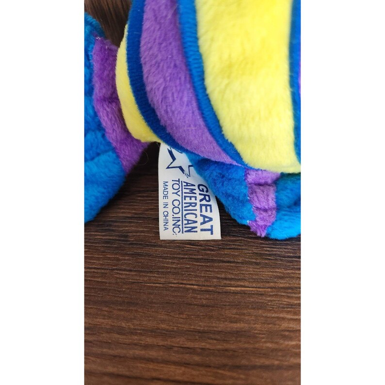 May include: A plush toy fish with yellow, purple, and blue stripes. The toy is labeled "Great American Toy Co., Inc." and "Made in China."