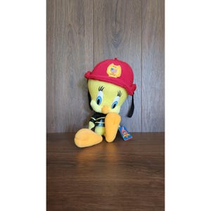 May include: A plush Tweety Bird toy wearing a red firefighter helmet with a gold emblem. The yellow bird is dressed in a black and yellow firefighter uniform. The toy has a tag attached.