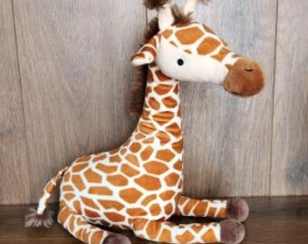 Könnte beinhalten: Ein Giraffen-Plüschtier mit cremefarbenem Gesicht und einem braun-weißen Fleckenmuster auf dem Körper. Das Stofftier hat zwei kleine braune Haarbüschel auf dem Kopf, dunkelbraune Hufe und einen langen Hals in sitzender Position.