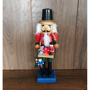 Peut inclure: Figurine de casse-noisette en bois avec un manteau rouge, des bottes noires et un grand chapeau noir. La figurine a une barbe et une moustache blanches et tient une balançoire avec des figurines miniatures. La base est bleue.