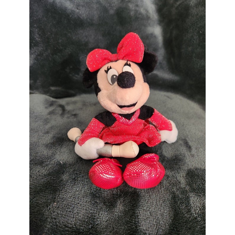 May include: A red and black plush toy of Minnie Mouse wearing a red sparkly dress and a red bow. She is holding a silver microphone.