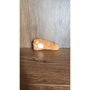 Puede incluir: Un juguete de cola de gato de felpa marrón claro con un diseño de huella de pata blanca. El juguete tiene una base negra y está sobre una superficie de madera. Para mascotas.