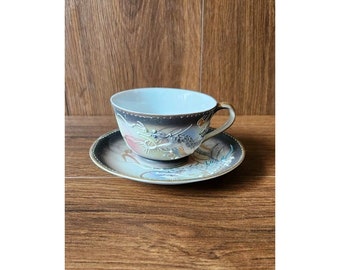 Könnte beinhalten: Eine weiße Teetasse und Untertasse mit einem schwarzen und goldenen Drachenmuster. Tasse und Untertasse sind mit einem bunten Blumenmuster verziert.