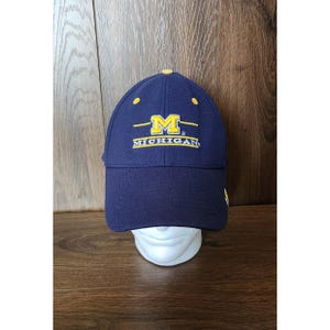 May include: A navy blue baseball cap featuring the yellow embroidered letter M and the word MICHIGAN in white text on the front panel. The cap has a yellow button on the top crown and structured stitching across the curved brim and panels.