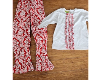 Op de afbeelding: Een rode en witte geprinte broek en een wit overhemd met lange mouwen met een rood ruchedetail. Het shirt heeft een groen label met het woord "Little" erop.