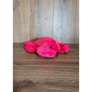 May include: A plush, hot pink lobster toy with black eyes and a black antenna. The toy is lying on a wooden surface. The lobster's claws and body are visible.
