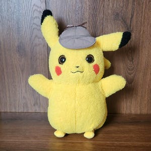 May include: A yellow plush toy character with large dark eyes, red circular cheeks, and black-tipped ears. The character wears a small, grey, brimmed detective-style hat with a brown ribbon detail on top. The plush has a soft, textured fabric.