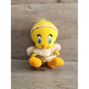 May include: A yellow plush Tweety Bird dressed as an angel with white wings and a gold halo. The plush toy is sitting on a wooden surface.