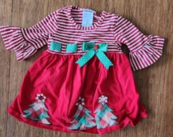 Könnte beinhalten: Ein langärmeliges Kleid mit rot-weiß gestreiftem Oberteil und gerüschten Ärmelabschlüssen. Die Taille ist mit einem grünen, gepunkteten Band und einer Schleife verziert. Der einfarbig rote Rock zeigt drei applizierte karierte Tannenbäume.