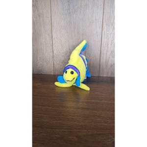 May include: A yellow plush toy shaped like a fish with blue and purple fins. The fish has a smiling face with black eyes.