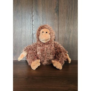 May include: A plush orangutan toy with brown fur, a tan face, hands, and feet. The stuffed animal is sitting on a wooden surface with a wooden background. The toy has a smiling expression.
