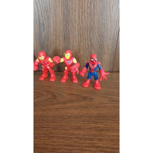 May include: Three superhero action figures: two Iron Man figures in red and gold, and a Spider-Man figure in red and blue. The figures are standing on a brown wooden surface.
