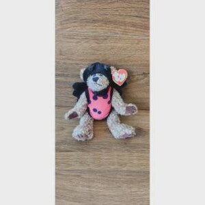 May include: A plush teddy bear with light brown fur wearing a black hood and a pink vest featuring dark circular spots. The bear has a small black nose and a heart-shaped red and white Ty tag attached to its ear, displaying the letters Ty.