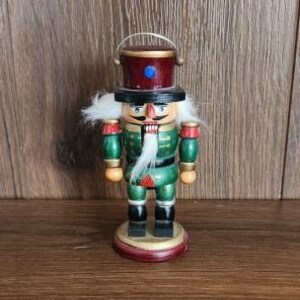 May include: A wooden nutcracker figurine featuring a red top hat with a blue gem, a green uniform with gold accents, black boots, and white synthetic hair. The figure has a painted face with a mustache and a white beard on a round base.