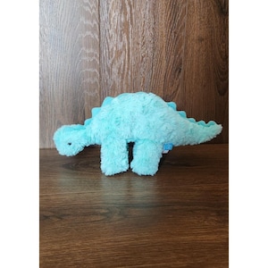 May include: A light turquoise plush dinosaur toy with a textured, fuzzy surface. The stuffed animal features a long neck, a small head with a single visible dark eye, and a row of soft, scalloped spikes running along its back and tail.