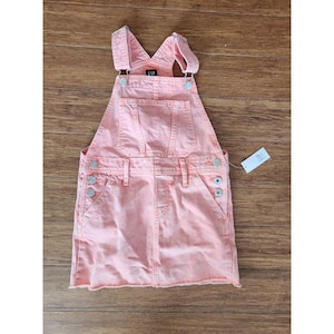 May include: A light coral denim overall dress with adjustable straps and silver-tone buttons. The dress features a front pocket and side pockets. The brand name "GAP" is visible on the bib. The hemline is frayed. A price tag is attached.