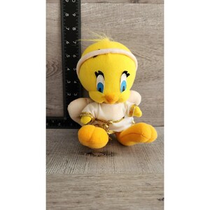 May include: A yellow plush Tweety Bird wearing a white angel costume with gold trim. The plush toy has blue eyes and a yellow beak.