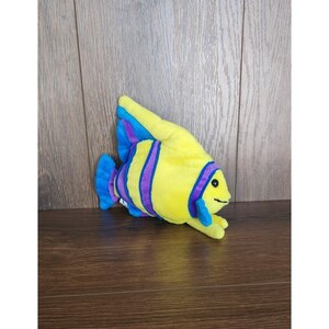 May include: Yellow plush fish toy with purple and blue stripes. The fish has a smiling face and a blue fin.